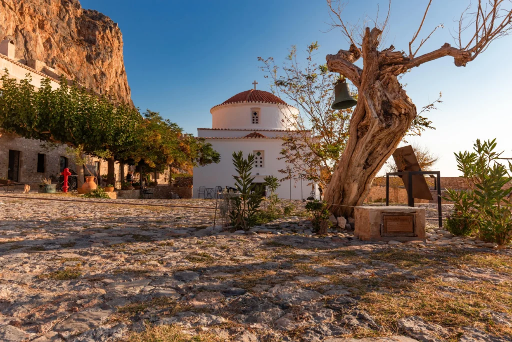 A view of a white church with a red-tiled dome, a rustic tree with a hanging bell, and a cobbled square in Monemvasia, Greece.