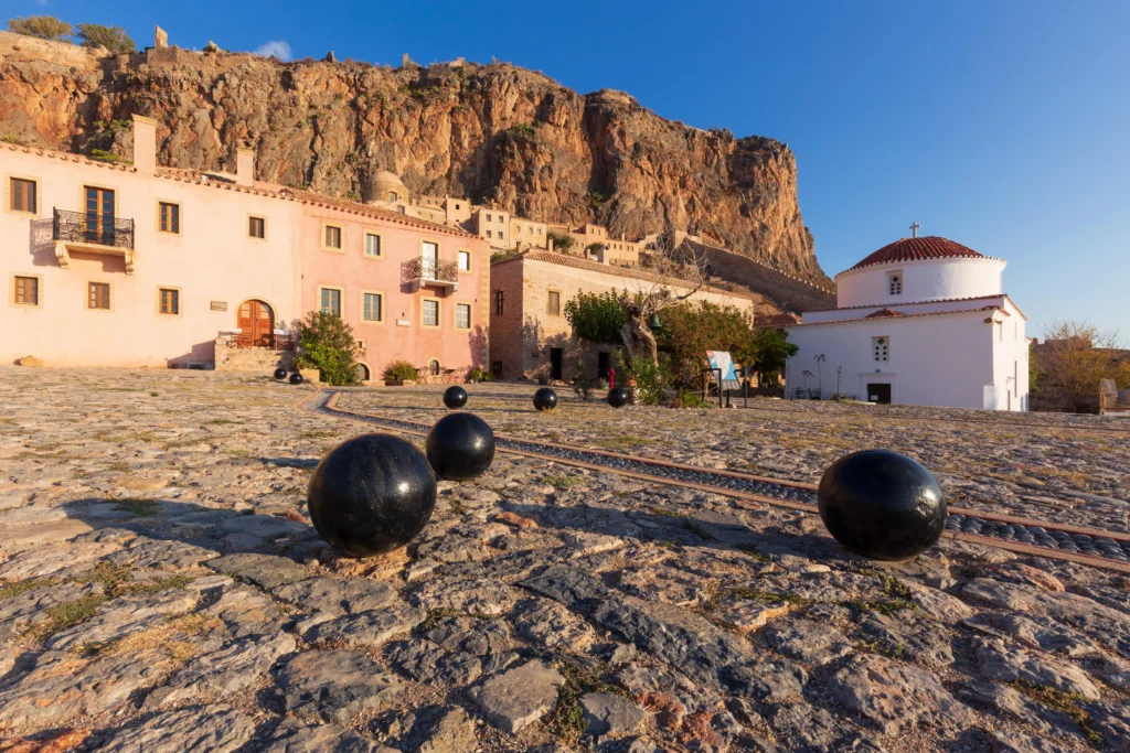 A cobbled square with black decorative balls in Monemvasia, featuring historic buildings, a white church with a red-tiled dome, and the massive rock face in the background.