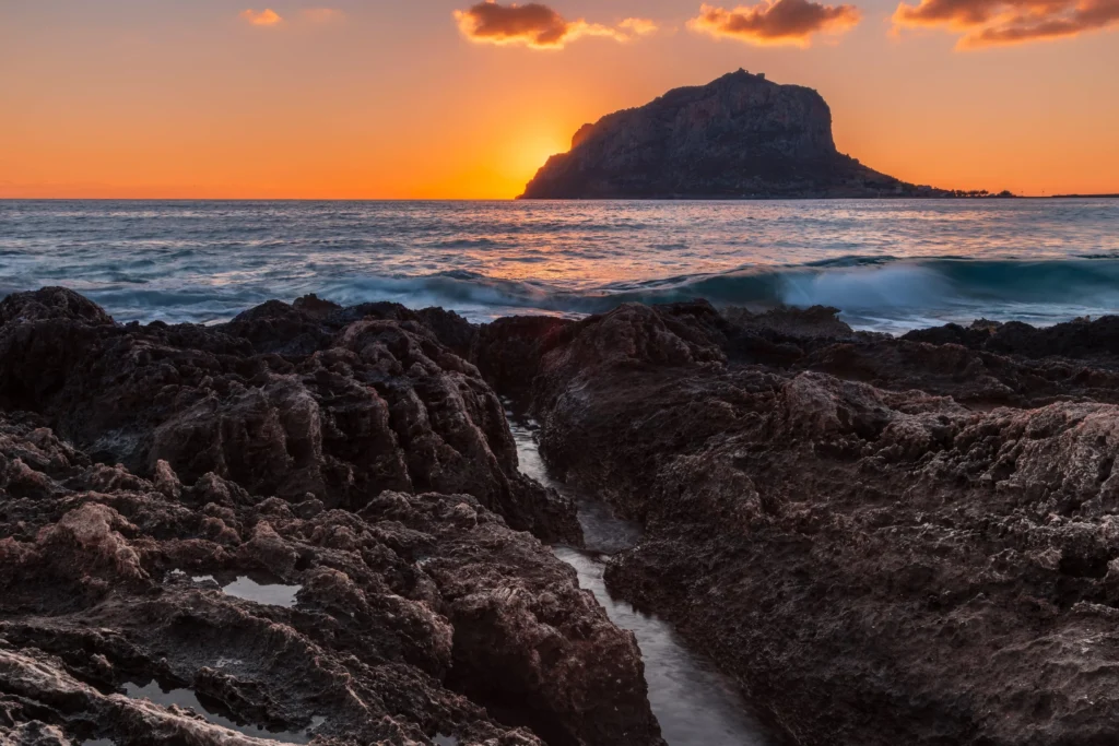 A colorful sunrise featuring jagged rocks in the foreground, gentle waves in the sea, and a giant rocky mountain island in Monemvasia, Greece.