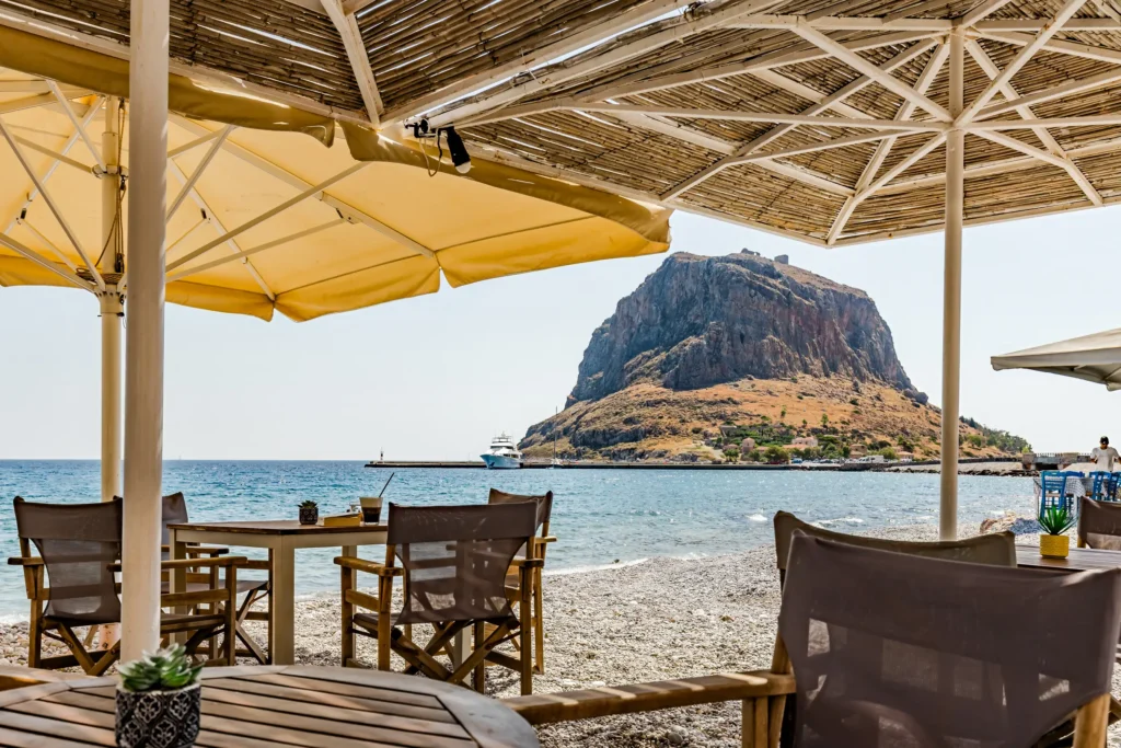 Beachside tables and chairs under umbrellas at Hotel Filoxenia overlooking the Monemvasia rock
